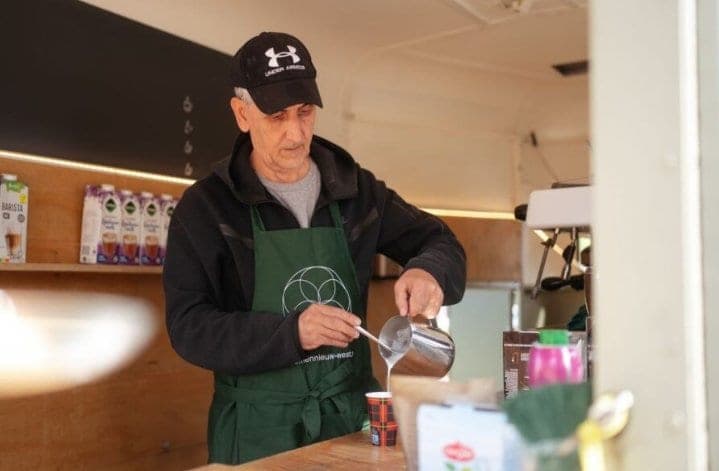 Een barista in een groen schort schenkt opgeschuimde melk in een koffiebeker in de Koffiekar