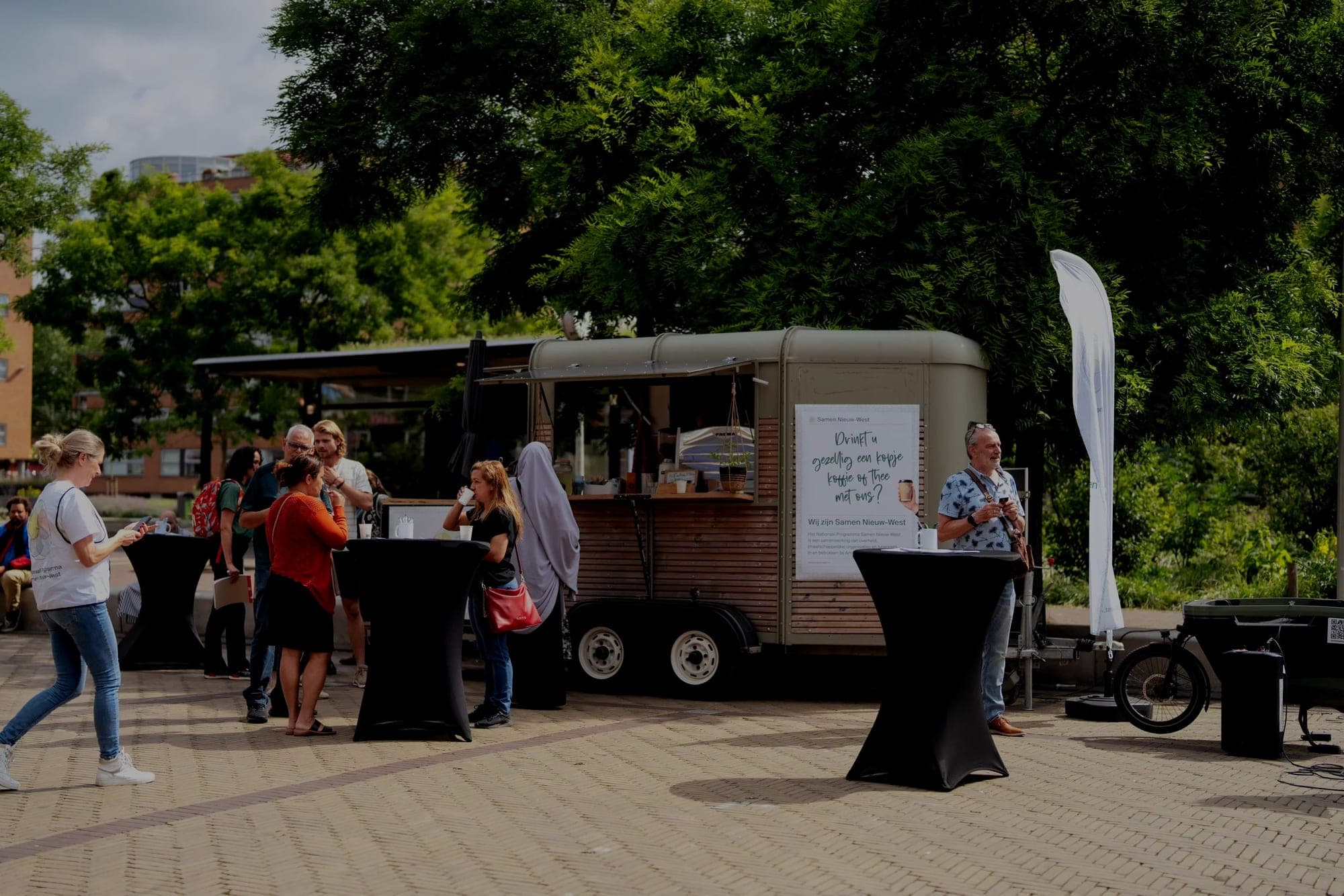 De groene Koffiekar van NEXUM op een plein in Nieuw-West, met bezoekers rond statafels onder grote bomen