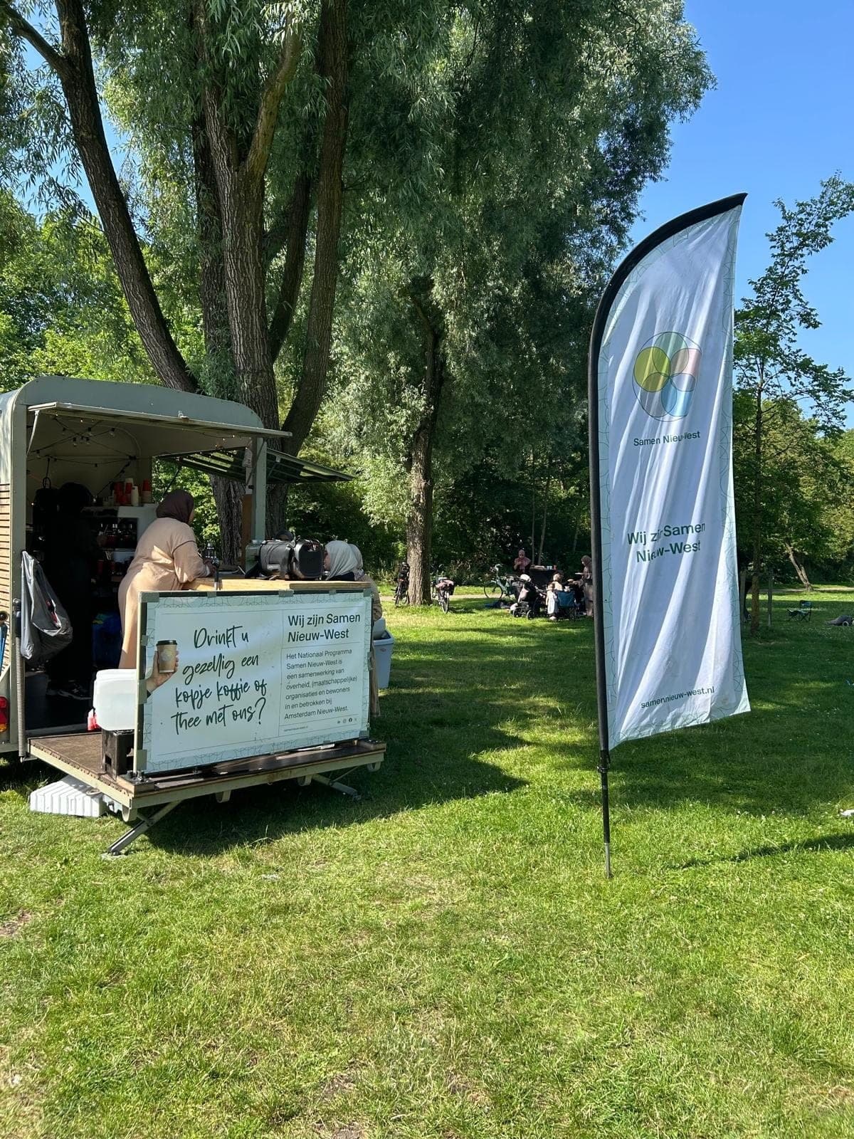 De Koffiekar in een zonnig park met een bord ‘Drinkt u gezellig een kopje koffie of thee met ons?’ en een vlag van Samen Nieuw-West
