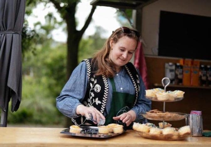 Een vrouw in een groen schort bestrooit cupcakes bij de Koffiekar, naast een etagère met koekjes
