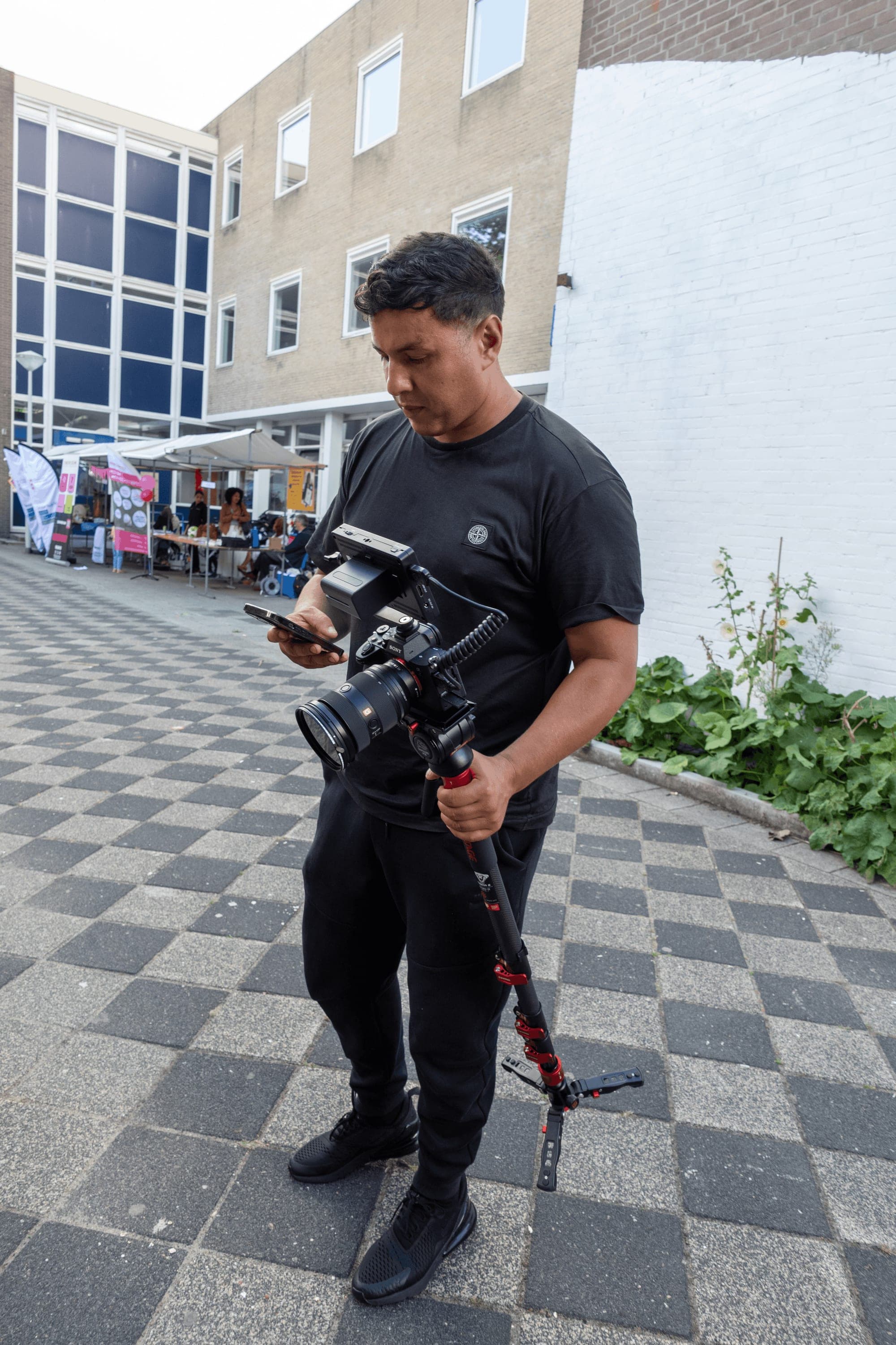 Een jonge videomaker van Y.O.U. met een camera op statief kijkt op zijn telefoon op een plein in Nieuw-West