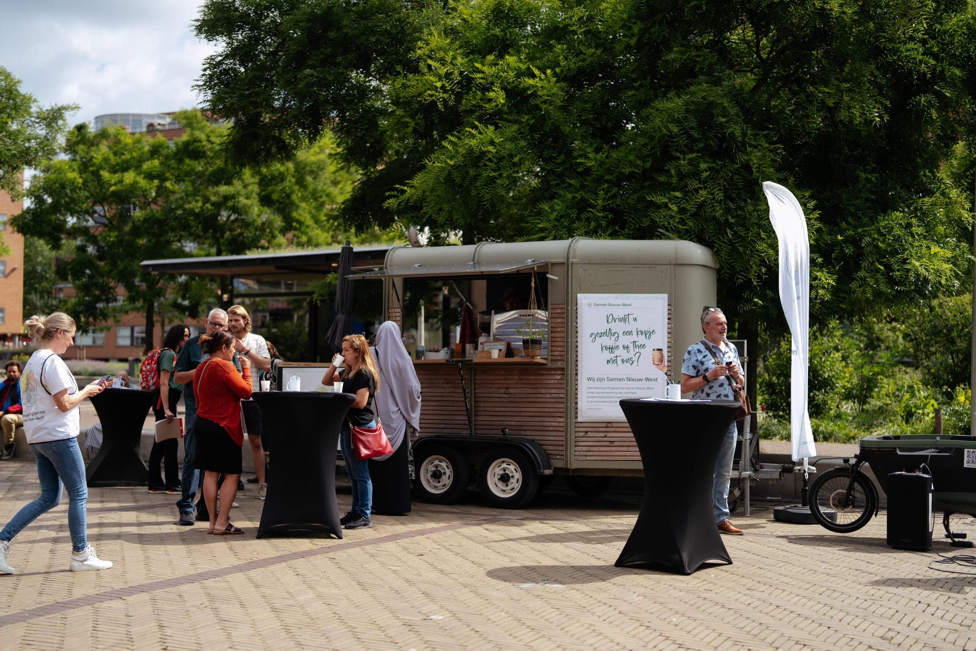 Bezoekers drinken koffie bij de groene Koffiekar op een zonnig plein; mensen staan aan zwarte statafels