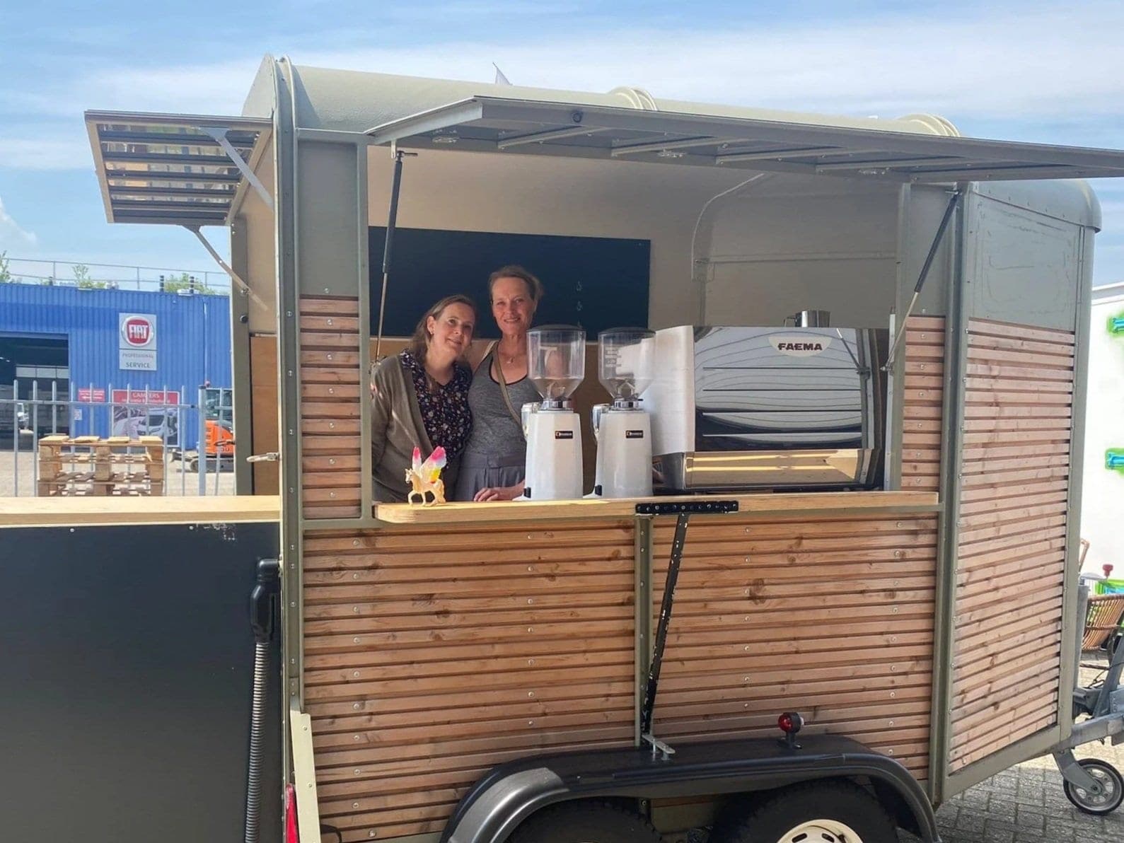 Twee vrouwen staan lachend achter de toonbank van de houten Koffiekar met espressomolens