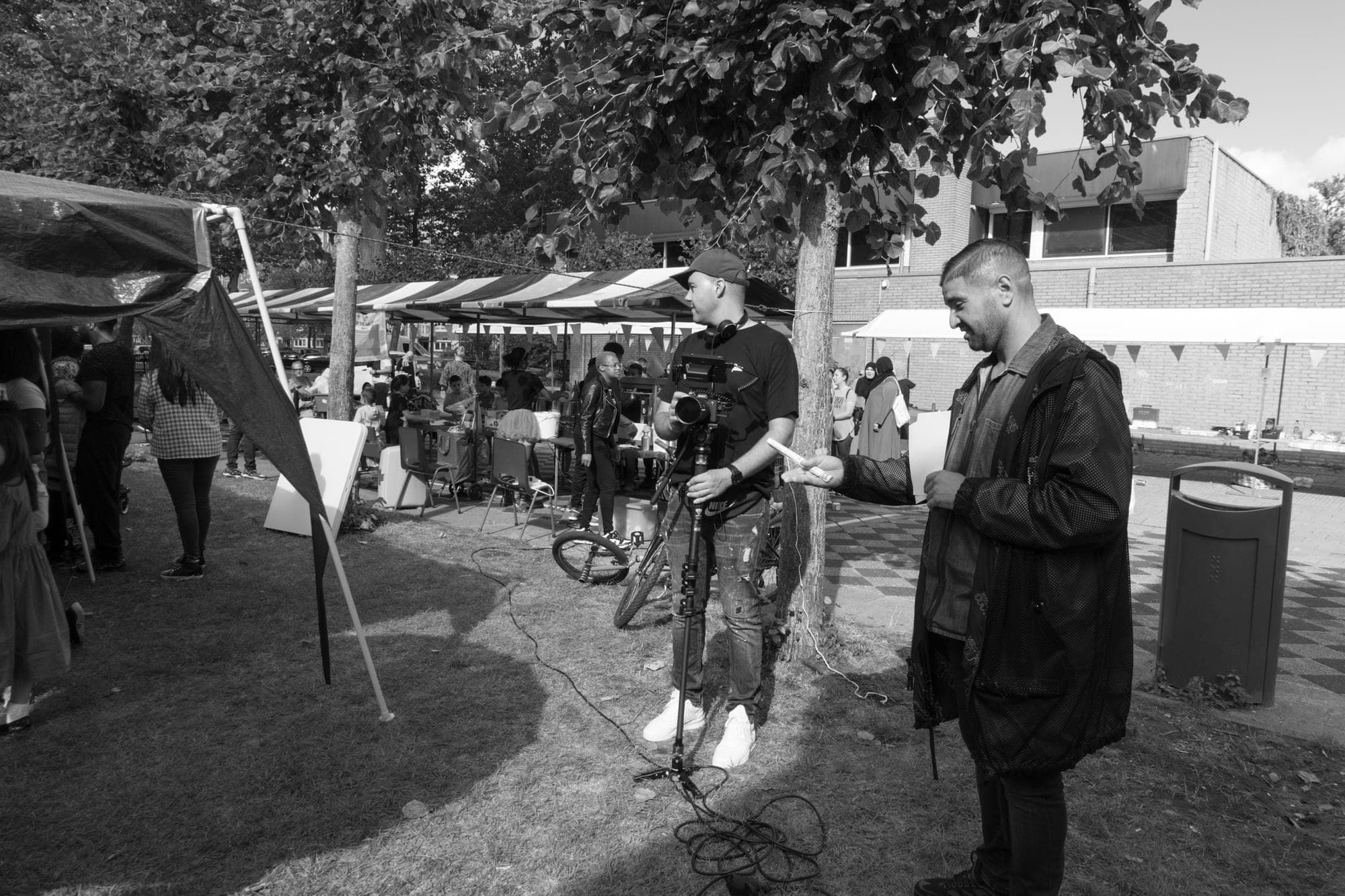 Zwart-witfoto van een filmopname op een buurtfeest: een cameraman met camera op statief en een man die een scène aanwijst